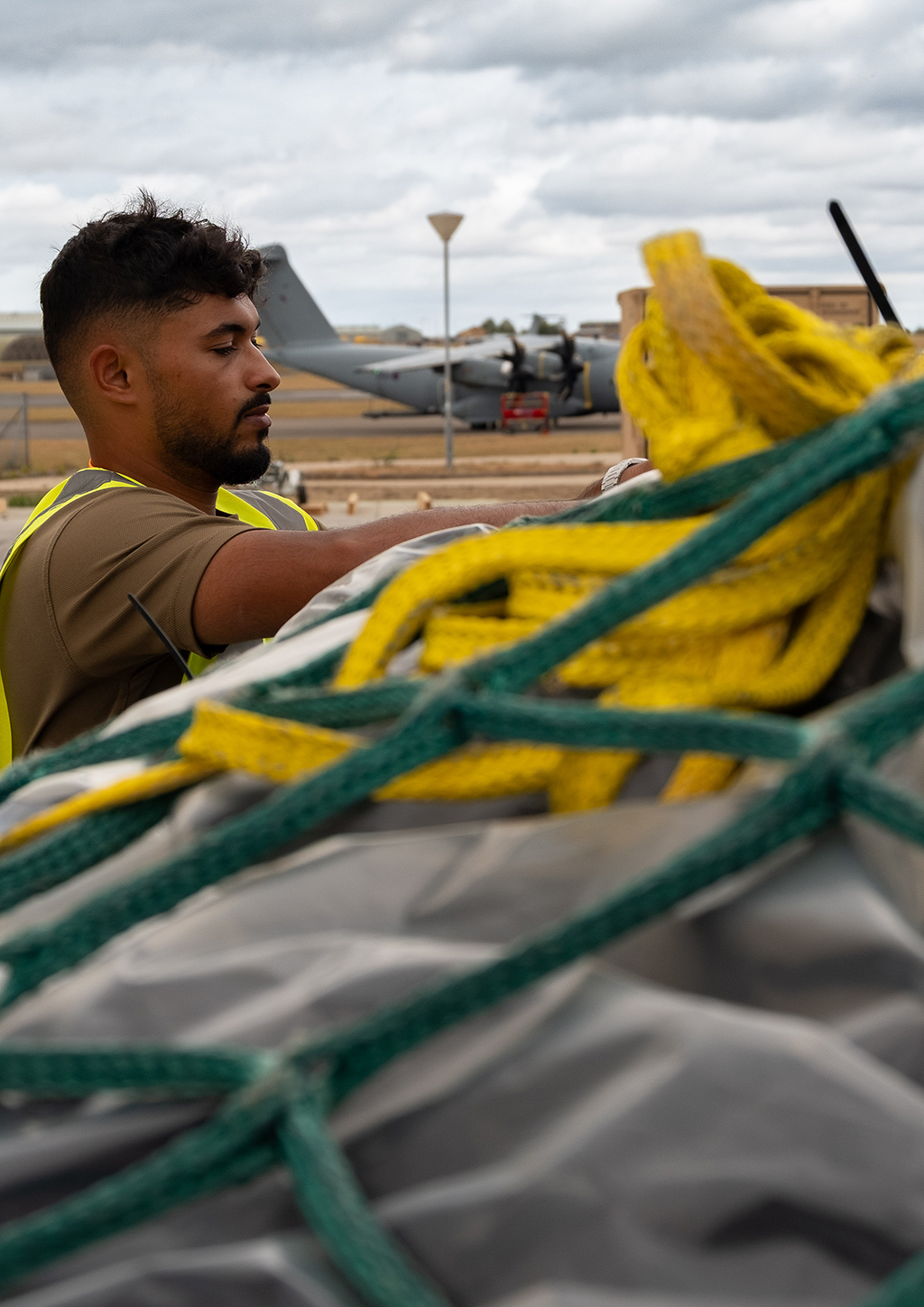 A member of 1 Air Mobility Wing, in a dark green t-shirt and a high-visibility vest, secures netting over cargo on a pallet on an airfield. The pallet is destined to be loaded onto a Atlas (A400M) aircraft.
